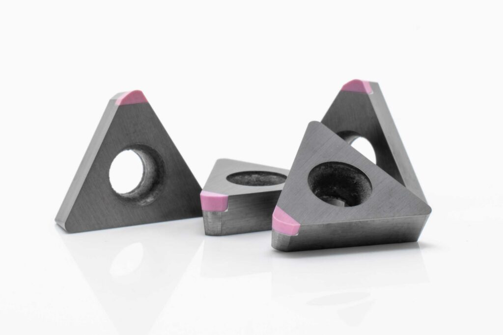 Drei metallene, dreieckige Körper mit runden Aussparungen und rosa Markierungen auf einer Seite, auf weißem Grund.