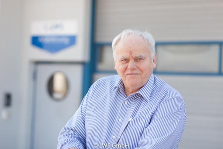 Älterer Herr in blau-weiß gestreiftem Hemd steht vor einem Unternehmenseingang.